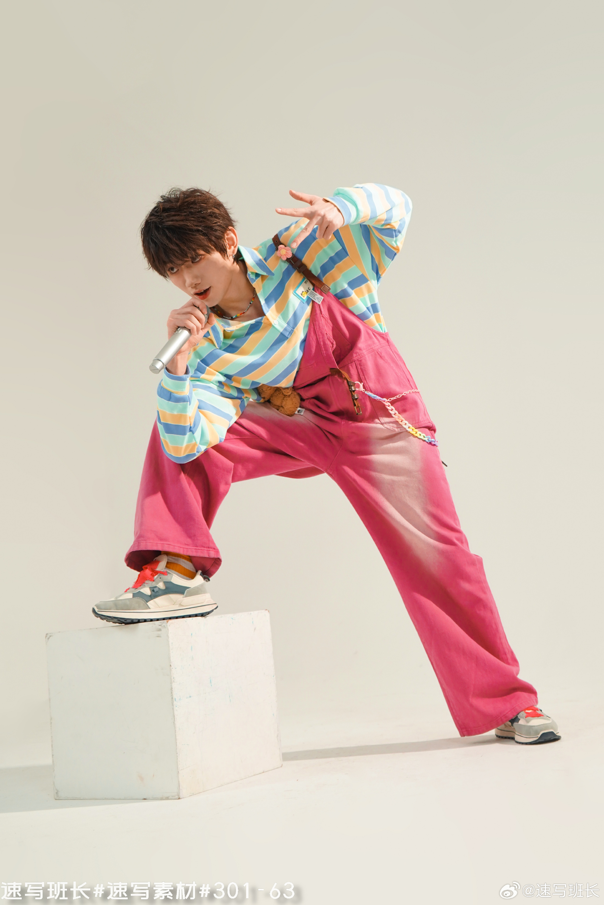 a young asian man dressed in a colorful, trendy hiphop outfit is posing with a microphone, as if he is singing. he is wearing a striped shirt, pink jumpsuit, and sneakers, and is standing on a white cube against a white background. the lighting is artificial and the image is taken from eye level.