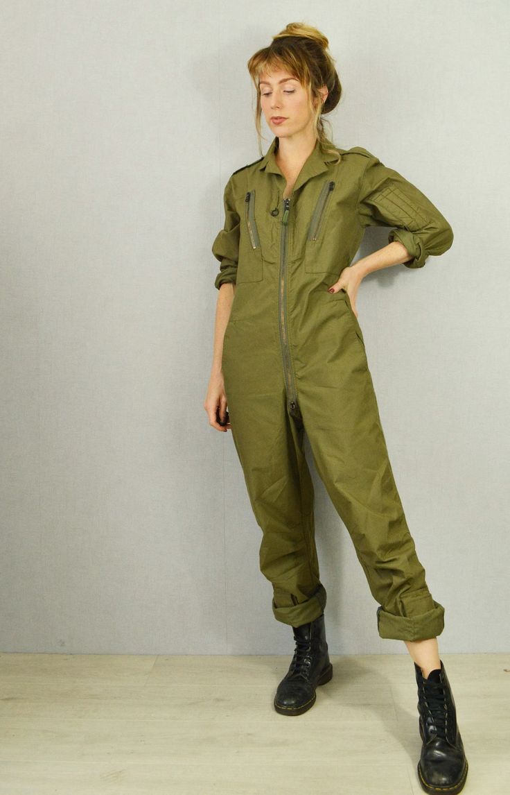 a fulllength shot of a woman wearing an olive green militarystyle jumpsuit. the jumpsuit has a zipper closure and multiple pockets. the woman is standing with her legs slightly apart and her hands near her sides.  she is wearing black doc martens boots and has her hair tied up in a bun. the background is a simple, plain grey.