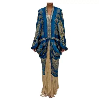 a person is wearing a long blue floral dress with tassels at the bottom. the dress appears to be a kaftan style. the background is white.