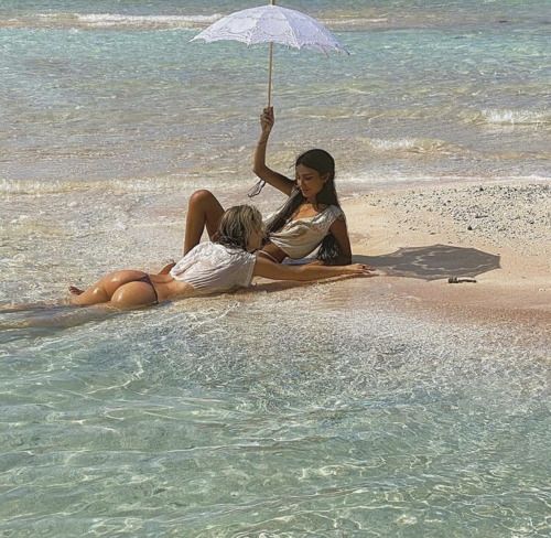 two women are relaxing on a beautiful beach, enjoying the sun and the clear blue water. they are wearing swimwear and are holding an umbrella. the sand is white and the ocean is calm and serene. this is a perfect place to relax and enjoy a tropical vacation.