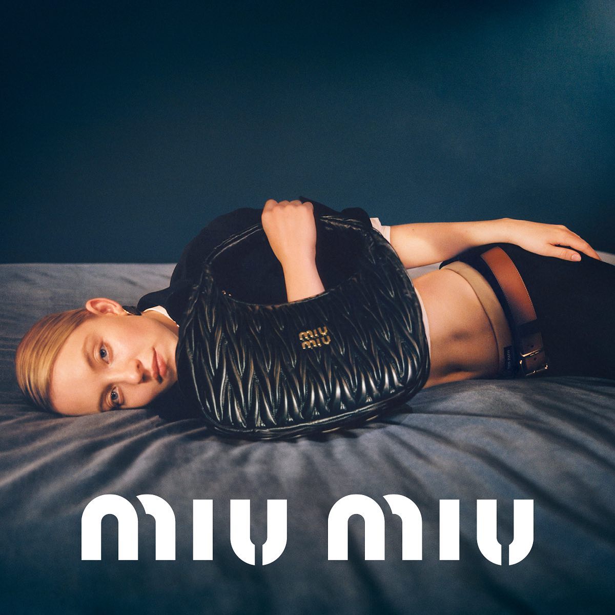 the image is a fashion advertisement for miu miu.  a young woman lies on a darkcolored bed, languidly holding a black quilted miu miu handbag. she's dressed in a dark top and pants, and her pose is relaxed and sensual. the background is a deep, moody bluegray. the miu miu logo is prominently displayed at the bottom of the image in a stylized white font. the overall aesthetic is sophisticated, minimalist, and suggestive of luxury.