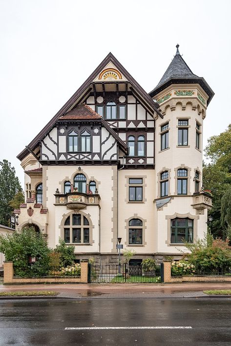 a photo of a large house with a turret on one side. it has multiple floors with many windows. the house has a small lawn and sidewalk in front and is located on a street. the sky is overcast.