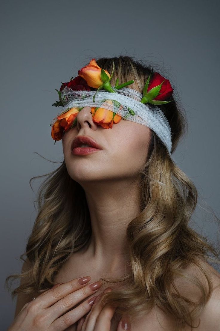 the image shows a closeup portrait of a young woman with long, wavy blonde hair.  her eyes are covered with a white bandage, which is adorned with several small, vibrant orange and red roses. the roses are arranged delicately across the bandage, partially obscuring her eyes. the woman has a serene expression, her hands gently clasped in front of her chest. she is bareshouldered, and the overall aesthetic is elegant, artistic, and slightly mysterious. the background is a simple, neutral gray, which draws attention to the model and the floral detail. the lighting is soft and flattering, enhancing the beauty of both the woman and the flowers.