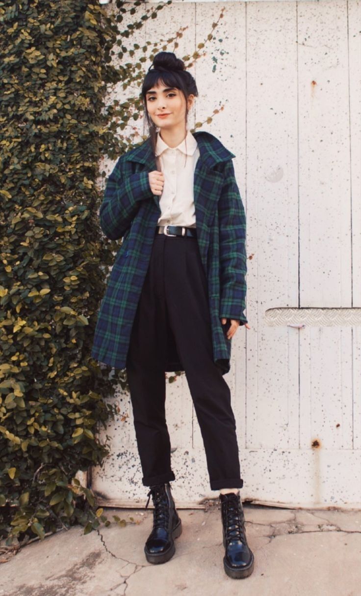 a young woman with dark hair styled in a bun and bangs stands against a white wall with ivy. she is wearing a green and black plaid coat over a creamcolored shirt, black highwaisted pants, and black dr. martens boots. she is looking slightly towards the camera with a relaxed expression.