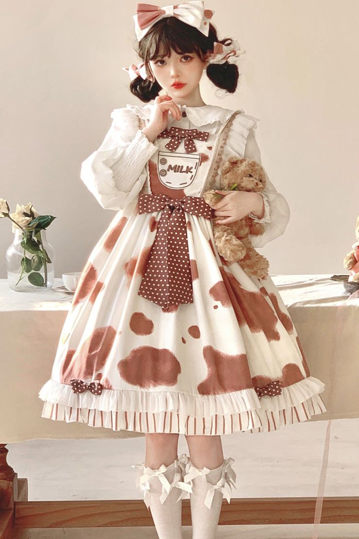 a young woman is posing in a kawaiiinspired outfit. she's wearing a white and brown cow print lolita dress with a milk carton design, a white blouse underneath, a matching headband, and white socks with brown ribbons. she's holding a brown teddy bear and looking at the camera.  the background features a white wall, a side table with a plant, and a blurred view of the rest of the room. the lighting is soft and natural.