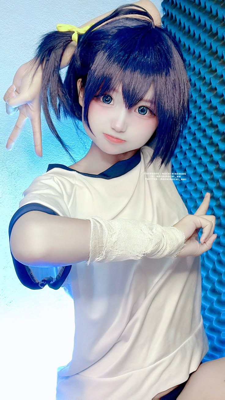a person in a cosplay outfit, likely an anime girl, is looking at the camera. the character has short, dark blue hair tied with a yellow ribbon, blue eyes, and pale skin. she is wearing a white uniform with blue trim and has a bandage wrapped around her left arm. her hands are making a peace sign. the background features a blue textured wall. the overall style suggests a cartoonish or animeinspired character brought to life through cosplay.
