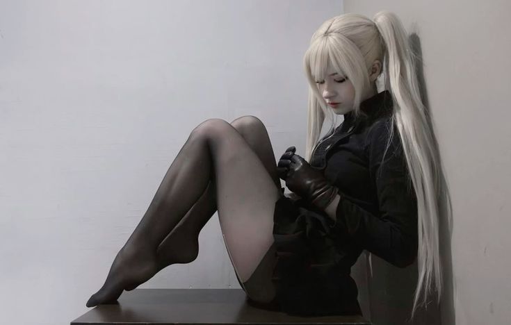 a woman wearing a blonde wig, black tights, gloves and a black dress is sitting. her hair is in pigtails.