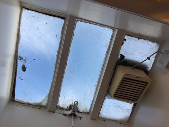 this is a lowangle view of three grimy windows. the windows are set into a white frame.  the top pane of each window is open. through the windows, we can see a bright blue sky with a few clouds.  a white ventilator fan is mounted on the wall to the right of the windows.  the white paint and window frames are showing signs of dirt and grime from age and use.