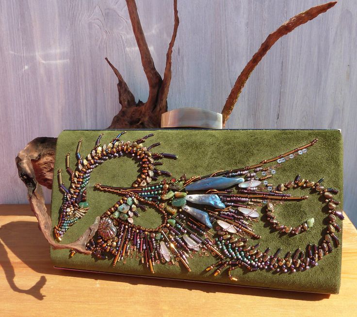 a closeup of a green clutch purse adorned with a beautifully beaded dragon design. the dragon is intricately crafted with various gemstones, beads, and sequins, creating a stunning and eyecatching piece. the clutch has a metallic clasp and is made of luxurious materials. the image is taken on a wooden surface, highlighting the bag's texture and craftsmanship.