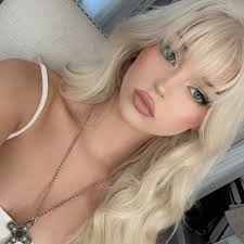 a selfie of a woman with blonde hair, bangs, and blue eyes. she is wearing a white top and a necklace with a cross pendant. she has pink lipstick and makeup on. she is posing for the camera.