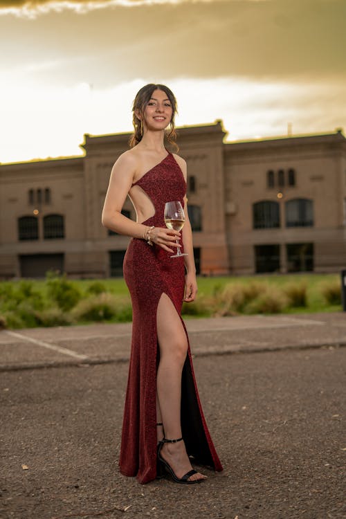 a young woman in a sparkling red oneshoulder evening gown with a high slit stands on a paved surface, holding a wine glass. she is wearing black heels and jewelry. in the background, there is a large building with windows and green vegetation. the sky is a warm, dusky color.