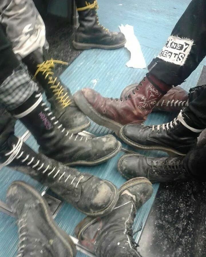 a gathering of people sitting or standing on a subway platform or vehicle, displaying their distinctive footwear. the ensemble includes a variety of doc martens boots, some with yellow laces and others with white, signifying different affiliations or subcultures within the punk or skinhead scene. the scene is characterized by the grunge aesthetic, showing worn and dirtied boots, which suggests a utilitarian and rebellious style. the focus on the footwear, combined with the backdrop, paints a picture of a subculture that blends fashion with elements of counterculture.