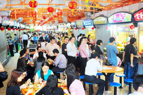he image shows a busy and vibrant hawker center in singapore.  numerous people are seated at tables, enjoying meals from various food stalls. the atmosphere is bustling and lively, with a mix of locals and tourists.  red lanterns and other chinese decorations hang from the ceiling, adding to the festive atmosphere.  the scene is full of color and activity, offering a glimpse into the everyday life and culinary culture of singapore.