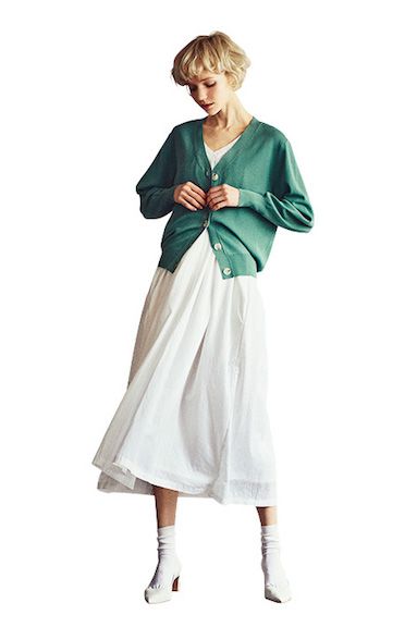 a woman is wearing a green cardigan and a white skirt with white heels. she is standing in a studio with a white background. her hair is a blonde bob. she is looking down and holding the cardigan closed.