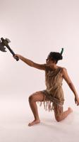 a woman in a fringed, tan outfit poses with an axe in a studio setting. the axe is raised in her right hand, and her left leg is extended behind her. she is looking past the axe. the background is a plain white, suggesting a studio environment. the lighting is soft and diffused, casting subtle shadows.