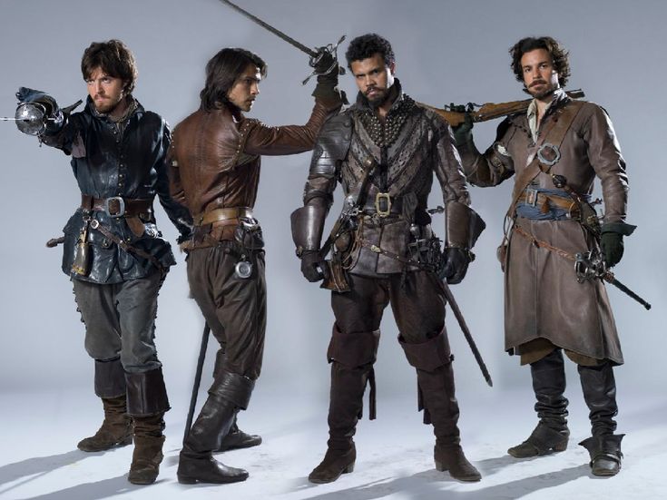 four men stand in front of a white background. they are all dressed in period costumes and they are all holding swords. the man on the far left is wearing a blue jacket and black pants. the second man is wearing a brown leather jacket and pants. the third man is wearing a brown leather jacket and pants with chainmail armor. the fourth man is wearing a brown leather coat and black pants.