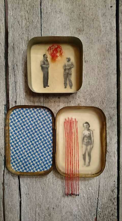 the image shows a mixed media art installation with two small, rectangular boxes placed on a weathered wooden background. the top box contains a vintage photograph of two men in uniform with a tangle of red thread suspended above them. the bottom box is open, revealing houndstooth fabric in the left section and a photograph of a woman in a swimsuit in the right section. red thread hangs vertically beside the woman.