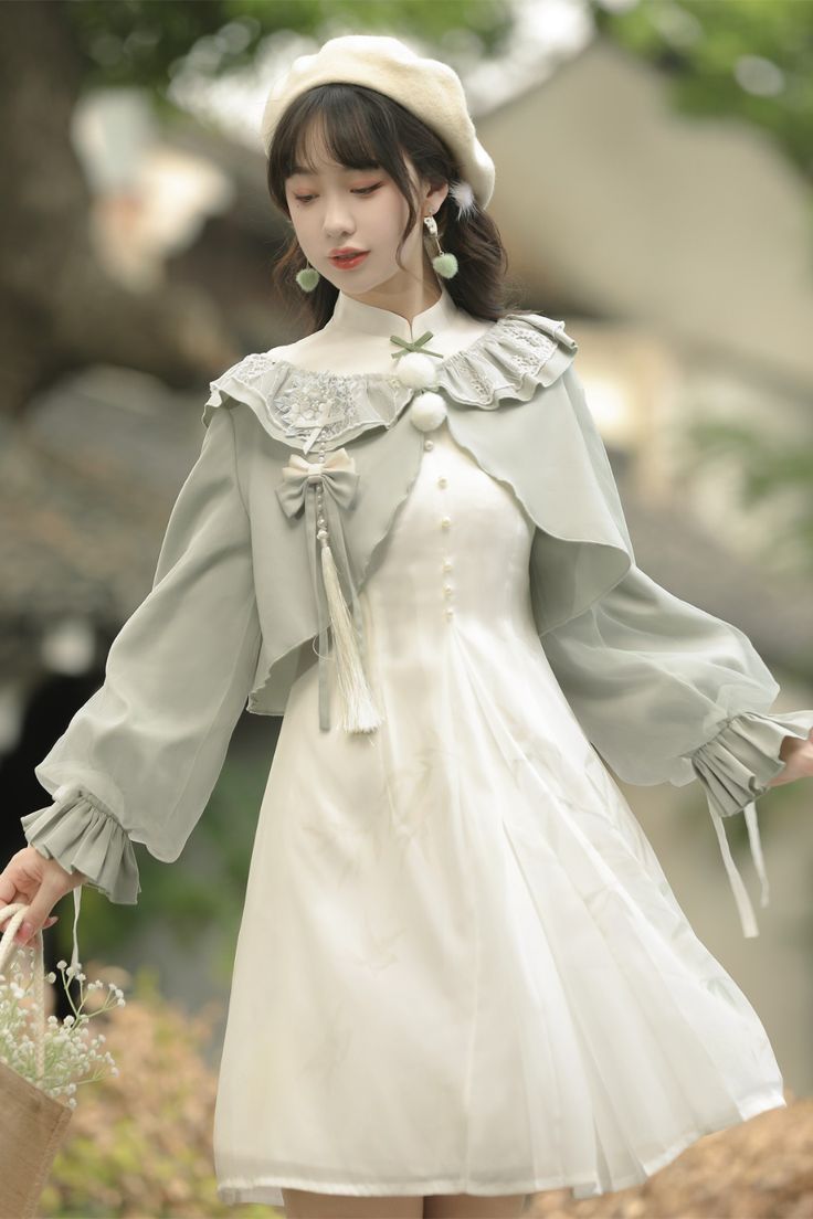 a young woman wearing a white dress with a light green jacket stands outside in a garden. the dress has a high collar and delicate lace detailing. the jacket is cropped and features long, flowing sleeves and decorative ties. the woman is also wearing a white beret and holding a small bag. the overall style of the outfit is reminiscent of both traditional chinese clothing and lolita fashion.  the image is welllit with natural light and taken from an eyelevel perspective.