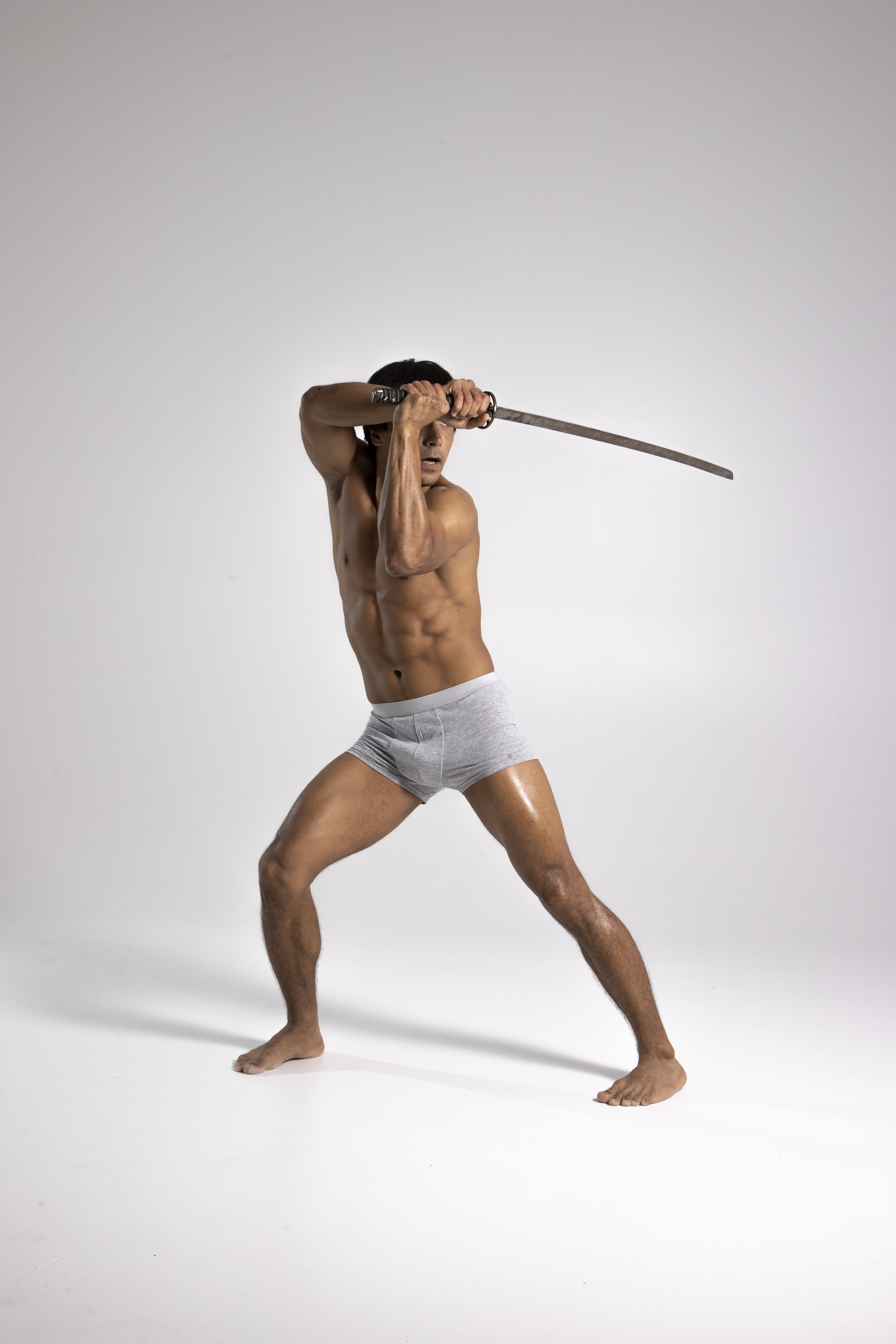 a muscular, shirtless man in gray shorts is holding a sword in a fighting stance in a studio shot against a white background. he is barefoot and appears athletic.