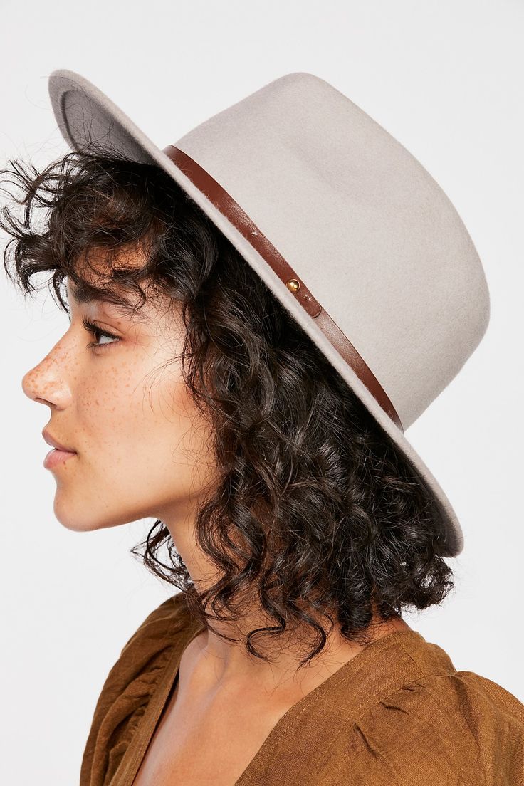 a woman with curly brown hair wears a gray fedora with a brown leather band. she is wearing a brown blouse and has a neutral makeup look.