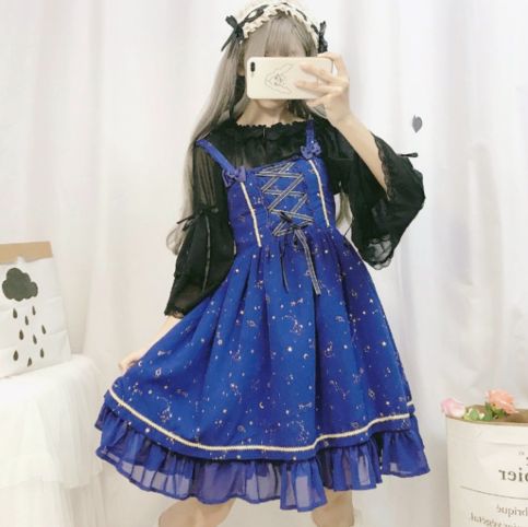 a young woman poses for a mirror selfie in a blue lolita dress with a celestial print. she has blonde hair with black accents and is wearing a black top underneath the dress. the dress has a laceup bodice, ruffled hemline, and white trim.  the background is white with a floral arrangement visible in the bottom right corner.