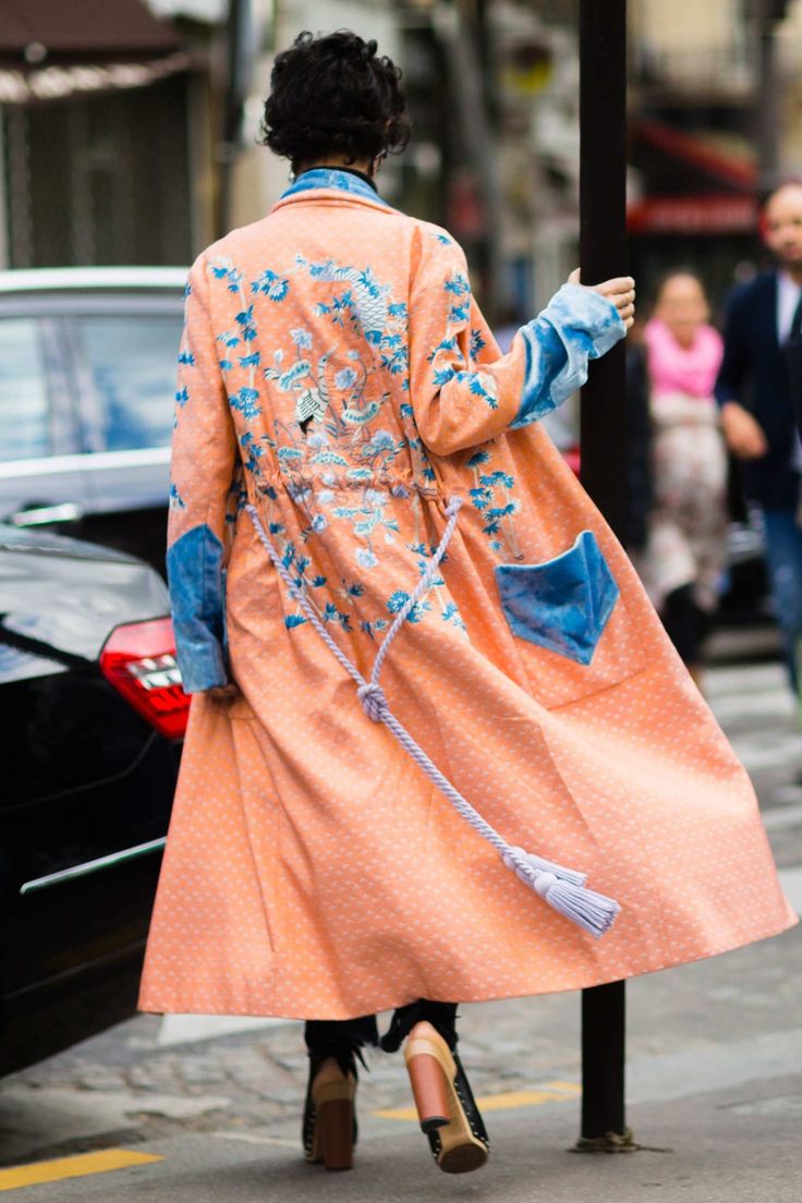 a woman wearing a long peach colored coat with blue denim details and floral embroidery walks down a city street. the coat is decorated with light lavender tassels and has rope detailing. she is wearing ankle boots and the photo is taken from behind.
