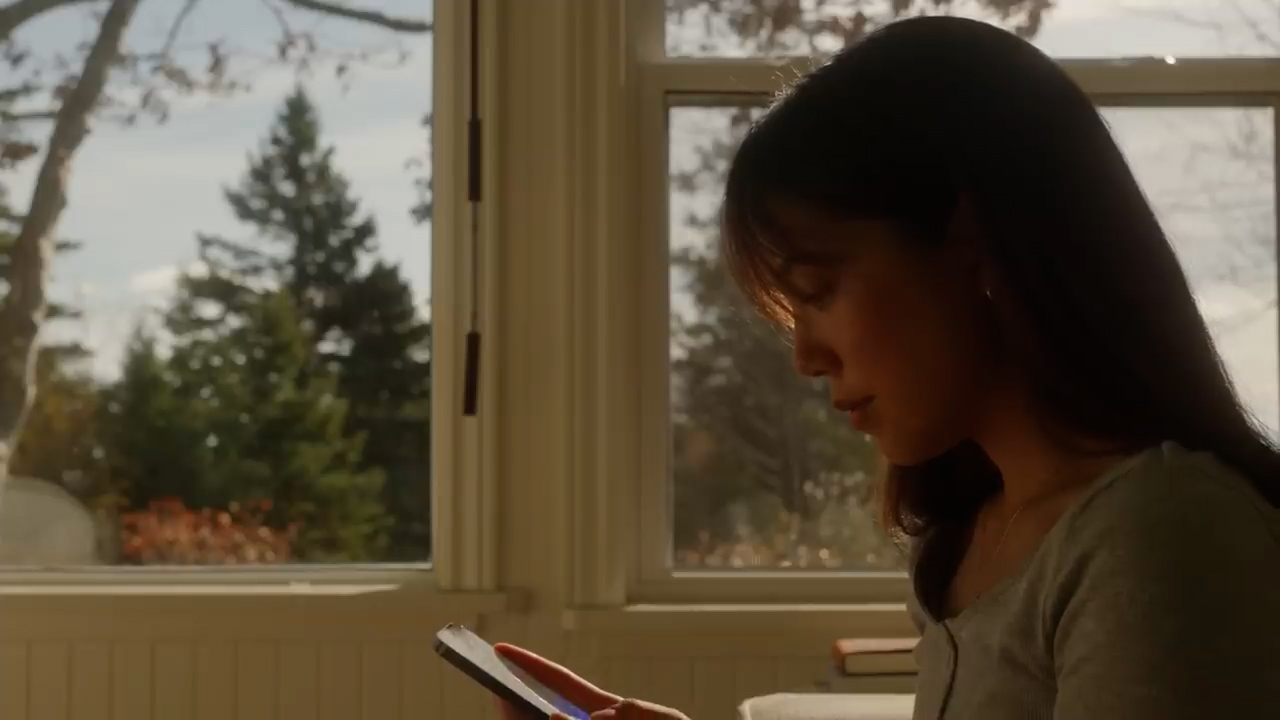 a woman with shoulder length dark hair is silhouetted against a brightly lit window. her face is in profile and angled downward towards the smartphone in her hands. the scene is warmly lit with sunlight streaming through the window behind her, revealing a view of trees and a glimpse of a blue sky. the overall ambiance suggests a quiet moment of contemplation.