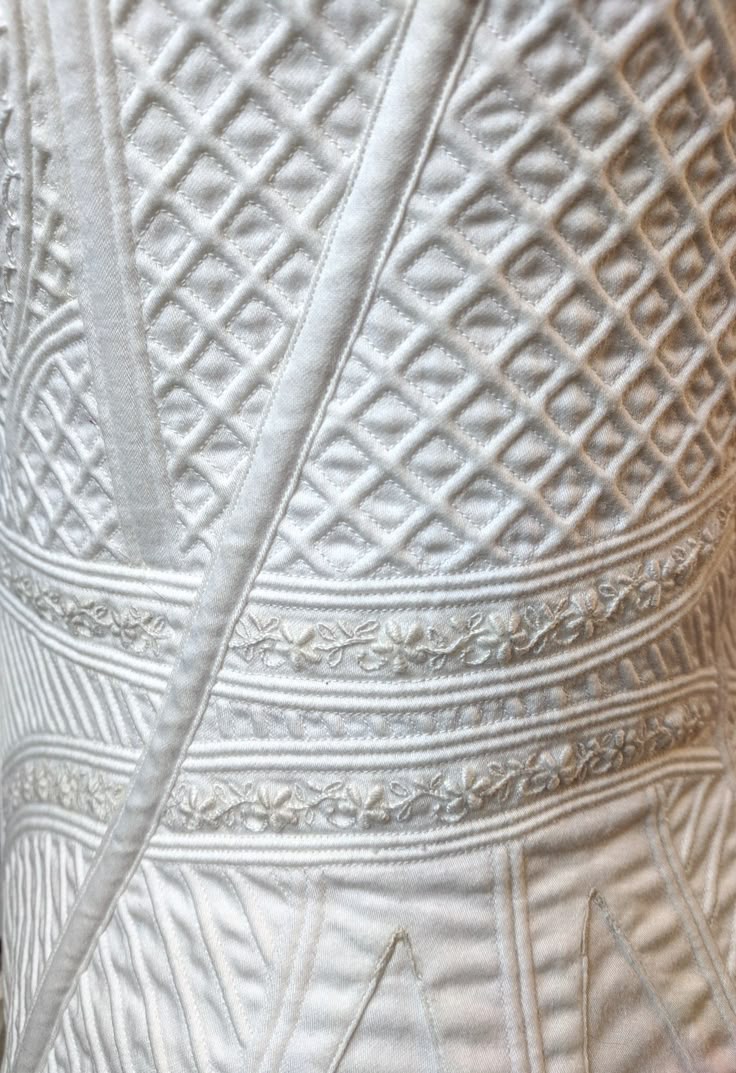 a closeup view of a white quilted fabric with a detailed diamond pattern on top. below the diamond pattern, there are horizontal lines with floral embroidery. a diagonal strip runs across the fabric. the texture appears soft and cushioned due to the padding.