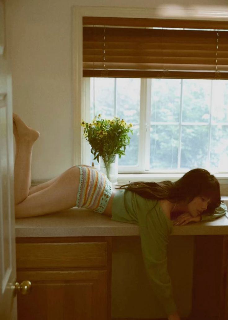a woman is lying on a kitchen counter. she is wearing multicolored crochet shorts and a green shirt. there is a vase of yellow flowers in front of a window with blinds behind her. cabinets are visible below the counter.
