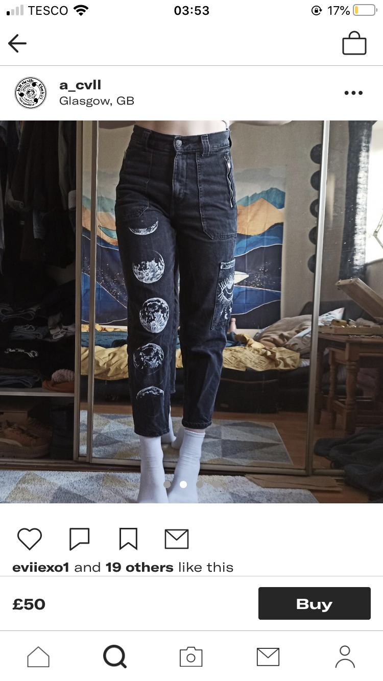 the image shows a pair of black denim jeans with a design of white moon phases painted down the front of the legs. the jeans are displayed on a mannequin or a person with their feet cropped out, set against a backdrop of a bedroom with a patterned tapestry. the image is welllit with natural light, giving the photo a bright and airy feel.