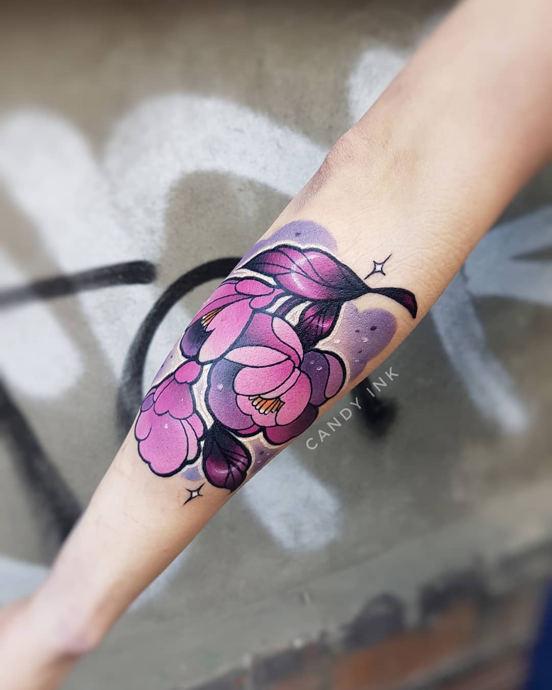 a closeup photo of a person's inner forearm, which features a colorful neotraditional tattoo of purple flowers with dark green leaves. the tattoo is surrounded by light purple shading and small white sparkles. the tattoo artist's signature, "candy ink," is visible in small black letters near the bottom of the tattoo. the photo is taken indoors under artificial lighting. the person's hand is not visible in the frame.
