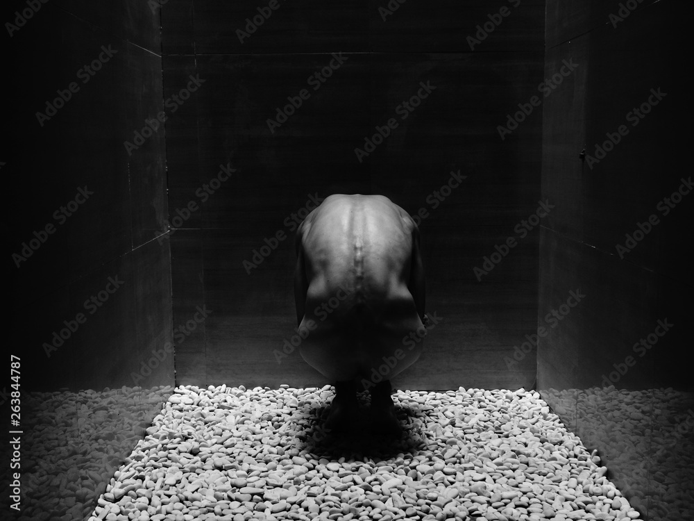a black and white studio shot shows a naked, kneeling woman in a room filled with stones. her back is to the camera. the walls are solid and dark with cracks showing their edges. the pebbles cover the floor creating a textured surface for the woman to kneel on. the lighting is dramatic, creating long shadows and highlights on the woman's body.