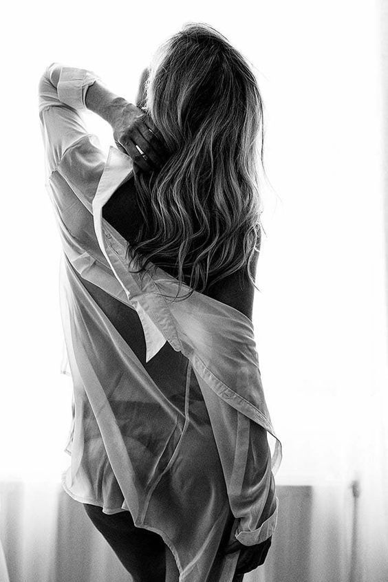 the image is a black and white photograph of a woman from behind. she has long, wavy hair and is wearing an oversized, sheer white shirt that is loosely draped over her body.  the shirt is partially open, revealing a glimpse of her back and shoulders. the lighting suggests a natural light source, likely from a window, creating a soft, intimate mood. the overall aesthetic is sensual and artistic.  the focus is on the woman's form and the way the fabric falls around her.