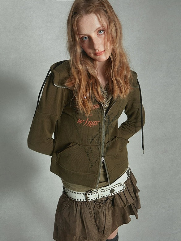 a young woman with long blonde wavy hair is modeling a casual outfit.  she's wearing an olive green zipup hoodie with some graphic design detail and a brown ruffled skirt cinched with a white studded belt. the setting appears to be a studio with a neutral gray backdrop.  the overall style is relaxed yet stylish, leaning towards grunge or alternative fashion.