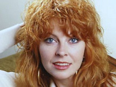 a closeup portrait of a young woman with long red hair. she is looking at the camera and smiling. she is wearing a white shirt and has a gentle, warm expression. she has bright blue eyes and a friendly smile.