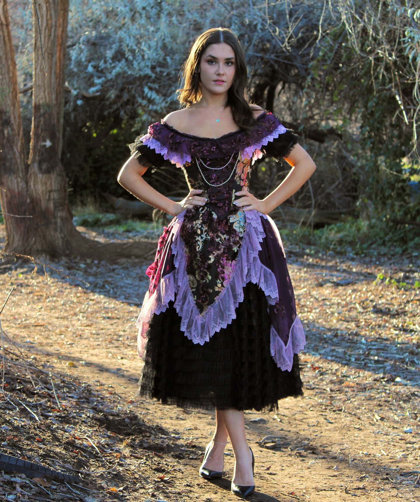 a young woman stands on a dirt path in a wooded area. she is wearing a long, elegant purple and black dress with lace detailing. the dress is styled like a vintage or historical gown. the woman is posing with her hands on her hips, and her expression is confident and pretty. the natural light and the surrounding nature create a picturesque scene.