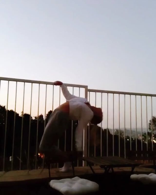 a woman is performing a yoga pose on a patio with a fence in front of a sunset sky. she is bending backward, holding onto the fence for support.