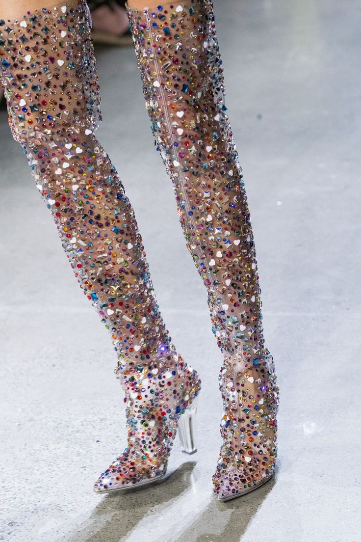 a model in a high fashion runway show wears a pair of clear, high heeled boots, that are covered in rhinestones. the boots are thigh high and are very sparkly and glamorous.