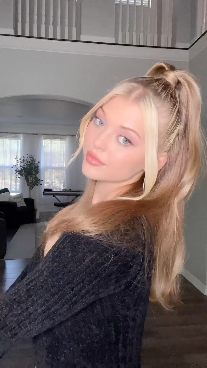 a woman with blonde hair in a high ponytail with faceframing pieces is posing in a room with white walls and a black sweater. the room appears to be a living room or common area with windows and a plant in the background.