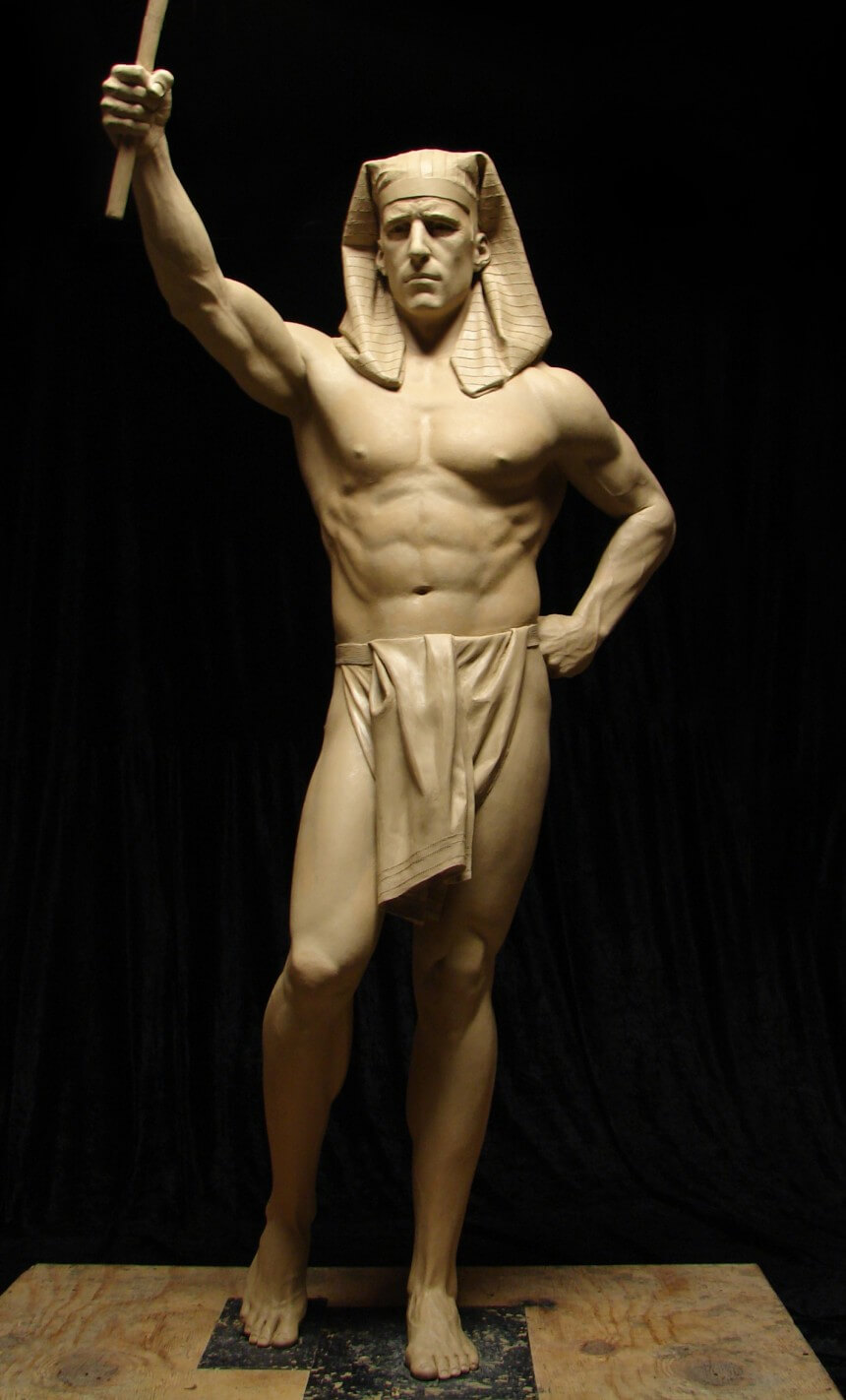 this image shows a clay sculpture of a man standing on a wooden platform. the sculpture depicts a muscular man of seemingly egyptian descent, as he is wearing a nemes headdress. he is holding a staff like object in his right hand, and is posed in a way that radiates power and authority. his left hand rests on his hip. the lighting is fairly dark, but illuminates the sculpture well. the background of the image is black. overall, the image exudes artistic talent and a passion for historical representation.