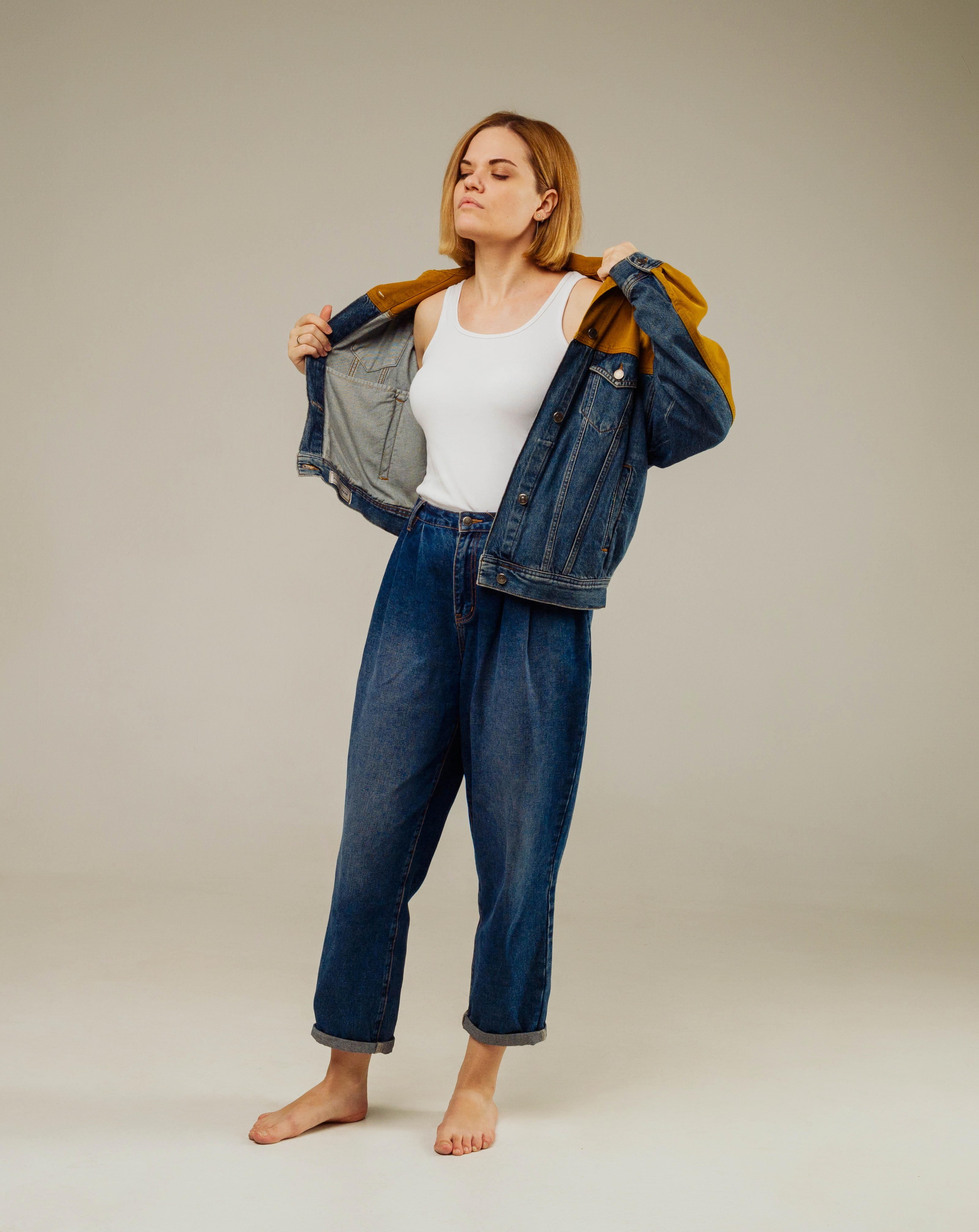 a young woman with short blonde hair stands barefoot on a gray background. she is wearing a white tank top, highwaisted blue jeans, and a denim jacket that she is holding open with both hands. her eyes are closed and she has a neutral expression on her face. the studio lighting is soft and even.