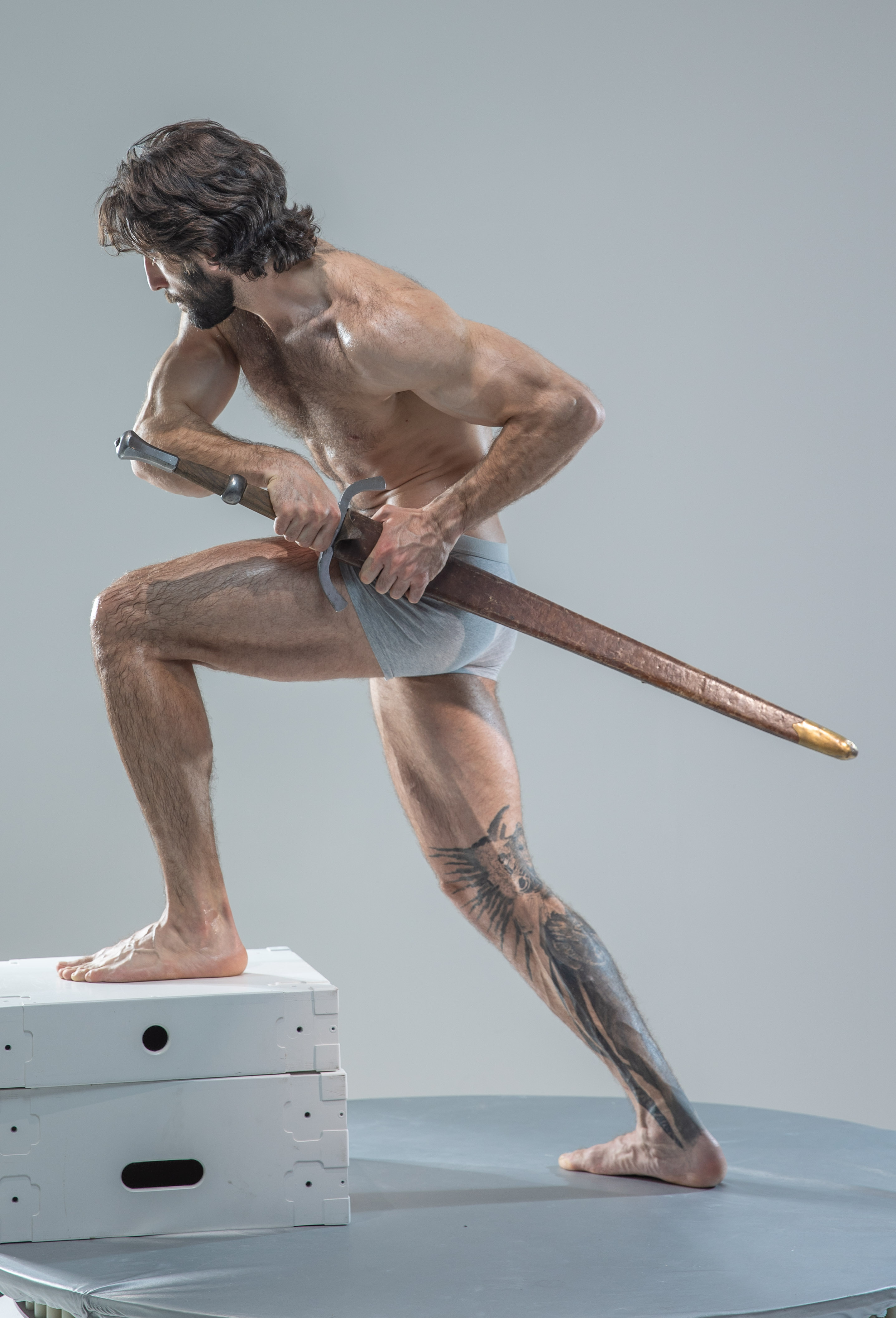 a muscular man with long dark hair and beard stands in a warriorlike pose. he holds a sword with both hands. he is shirtless, wearing only white underwear, which accentuates his physique. his right leg is bent, resting on a stack of white boxes, while his left leg is extended behind him. he has a large tattoo of a winged creature on his left calf. the background is a plain gray studio backdrop. he is illuminated by soft lighting that highlights his muscles and the curve of the sword.