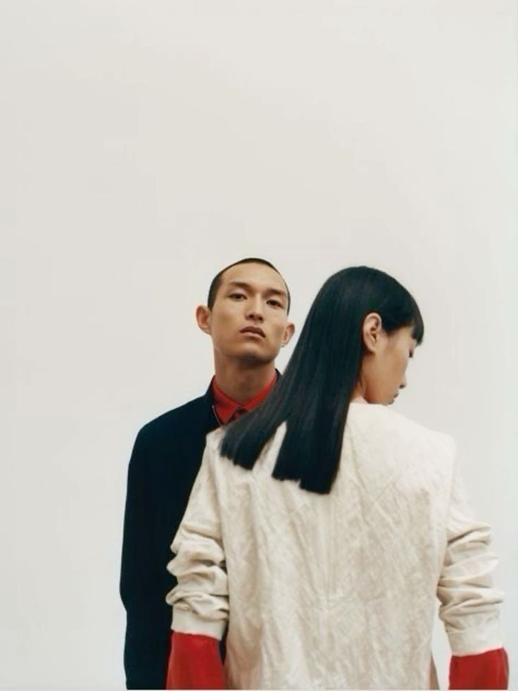 a couple posing in a studio. the man is wearing a black jacket and red shirt, while the woman is wearing a cream sweater. both are looking at the camera. the background is white.