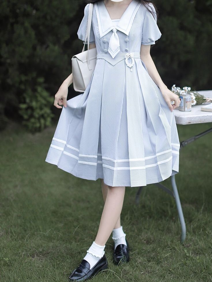 a young woman in a blue and white striped dress is standing in a garden. she is wearing white socks and black shoes and is holding the bottom of her dress. there is a white bag hanging on her left arm. the dress has a sailor collar and a white bow tie. she is posing for the camera. there is a picnic table in the background. the image is brightly lit and has a natural, outdoor setting.