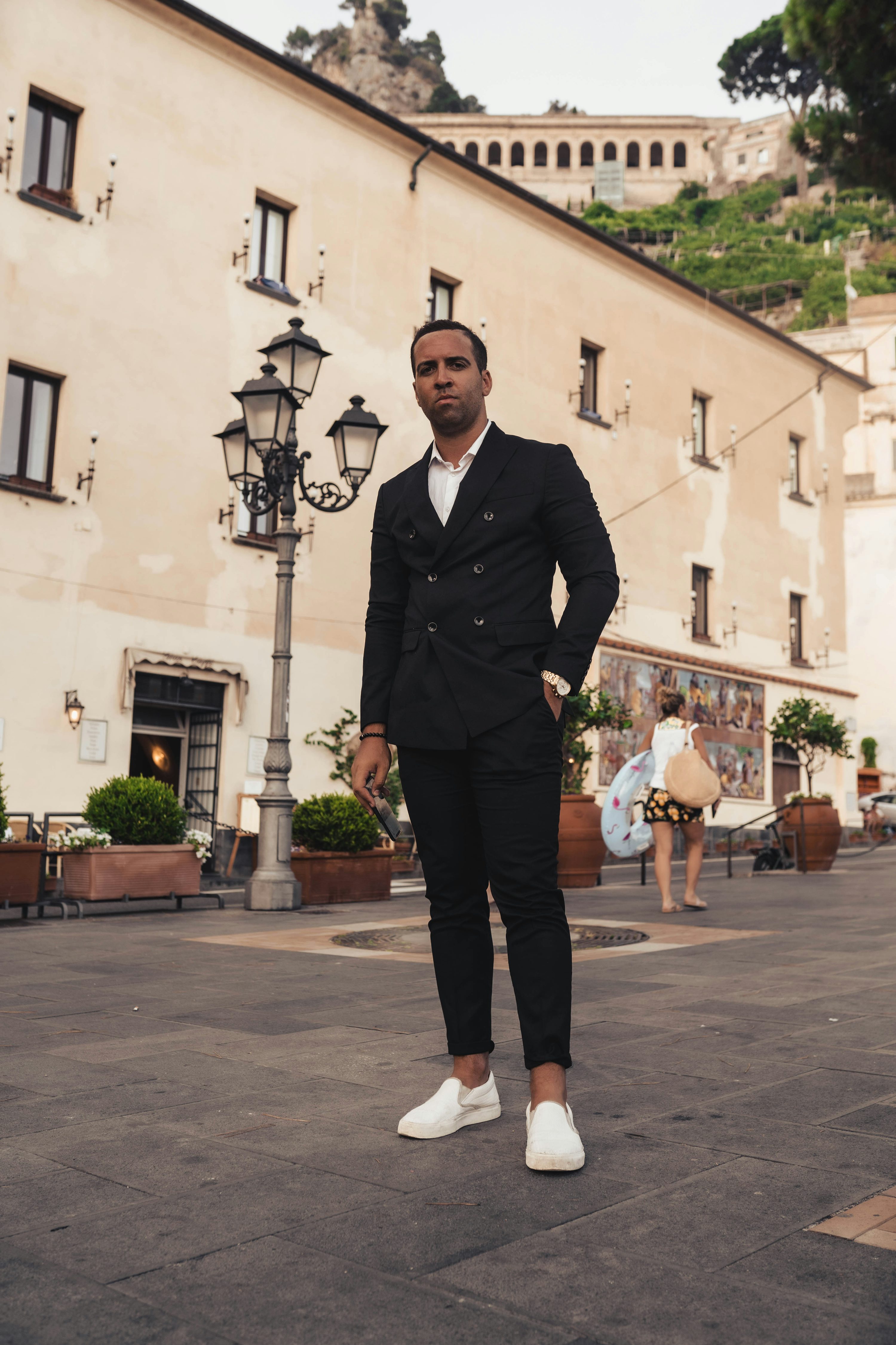 a welldressed man in a black doublebreasted suit and white slipon shoes stands confidently on a cobblestone street in a charming italian town.  the background features traditional architecture, a lamppost, and glimpses of other people in the distance. the setting appears to be sunny and touristy. the man's expression is serious and his posture suggests assurance.