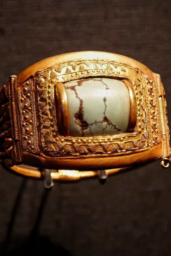 a closeup shot of an ornate gold bracelet on display in a museum. the bracelet is wide and features intricate detailing along its surface, including a central rectangular frame housing a large, pale green gemstone.  the background is dark and unfocused, drawing attention to the gleaming gold and delicate craftsmanship of the bracelet.