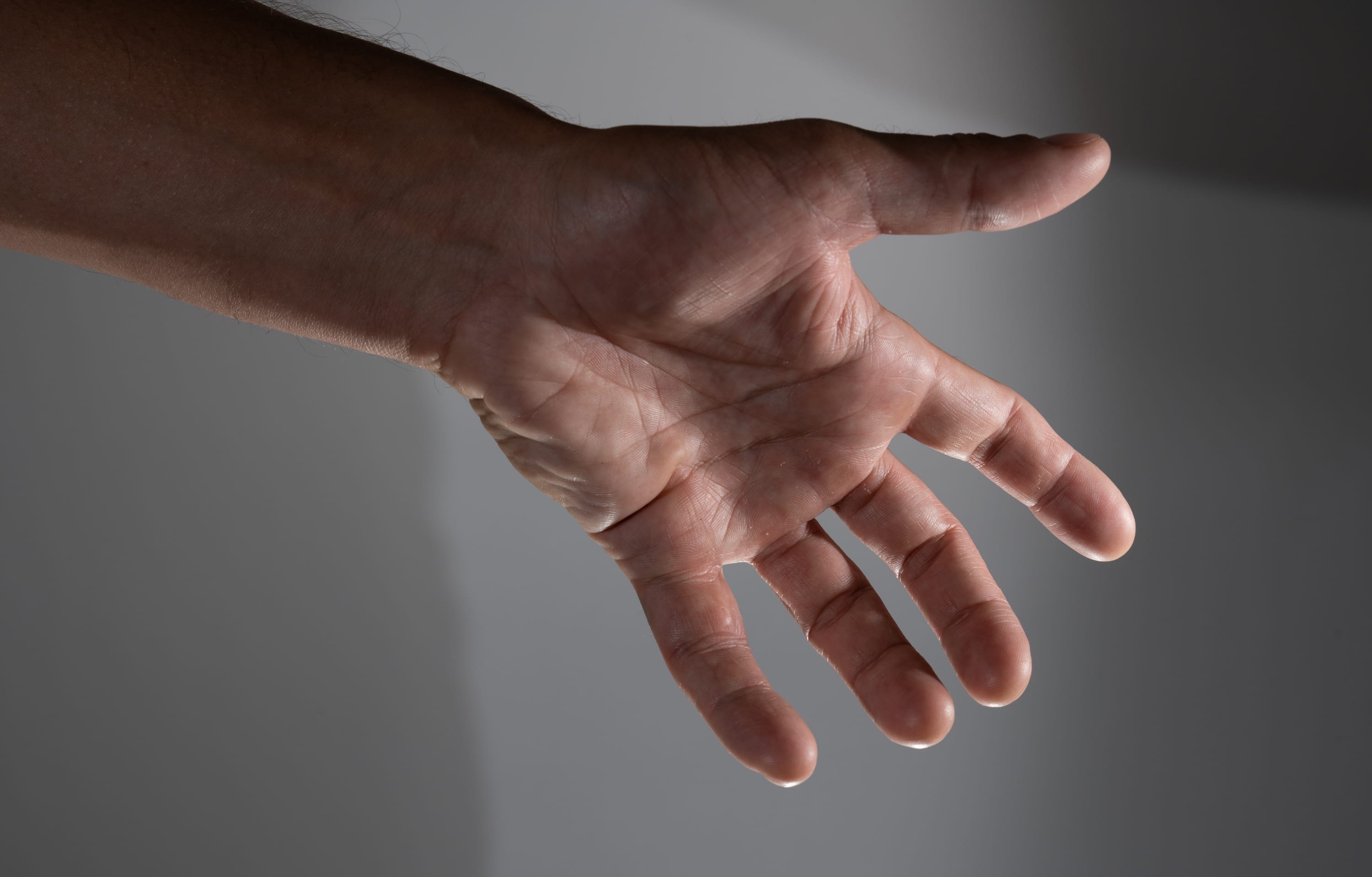 a closeup image of a man's outstretched hand against a gray background, illuminated by studio lighting. the image focuses on the details of the hand, including the wrinkles and lines on the palm and fingers. the hand is positioned as if reaching out or gesturing towards something.