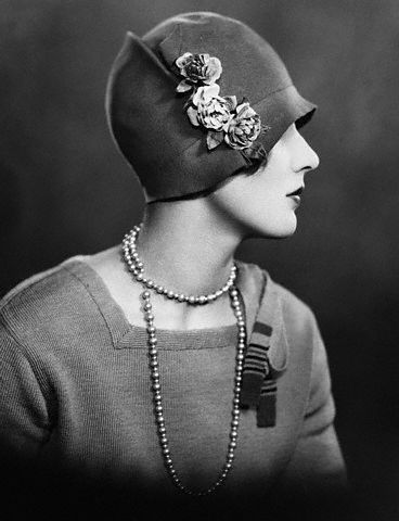a woman in a black and white portrait wearing a cloche hat with flowers and a string of pearls. she is looking slightly off to the left side of the image and appears to be elegant and sophisticated. the photo has a vintage feel to it.