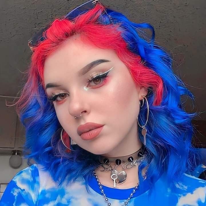 a portrait of a young woman with red and blue hair, wearing makeup, jewelry, and a tiedye shirt. she has multiple piercings and is looking directly at the camera.