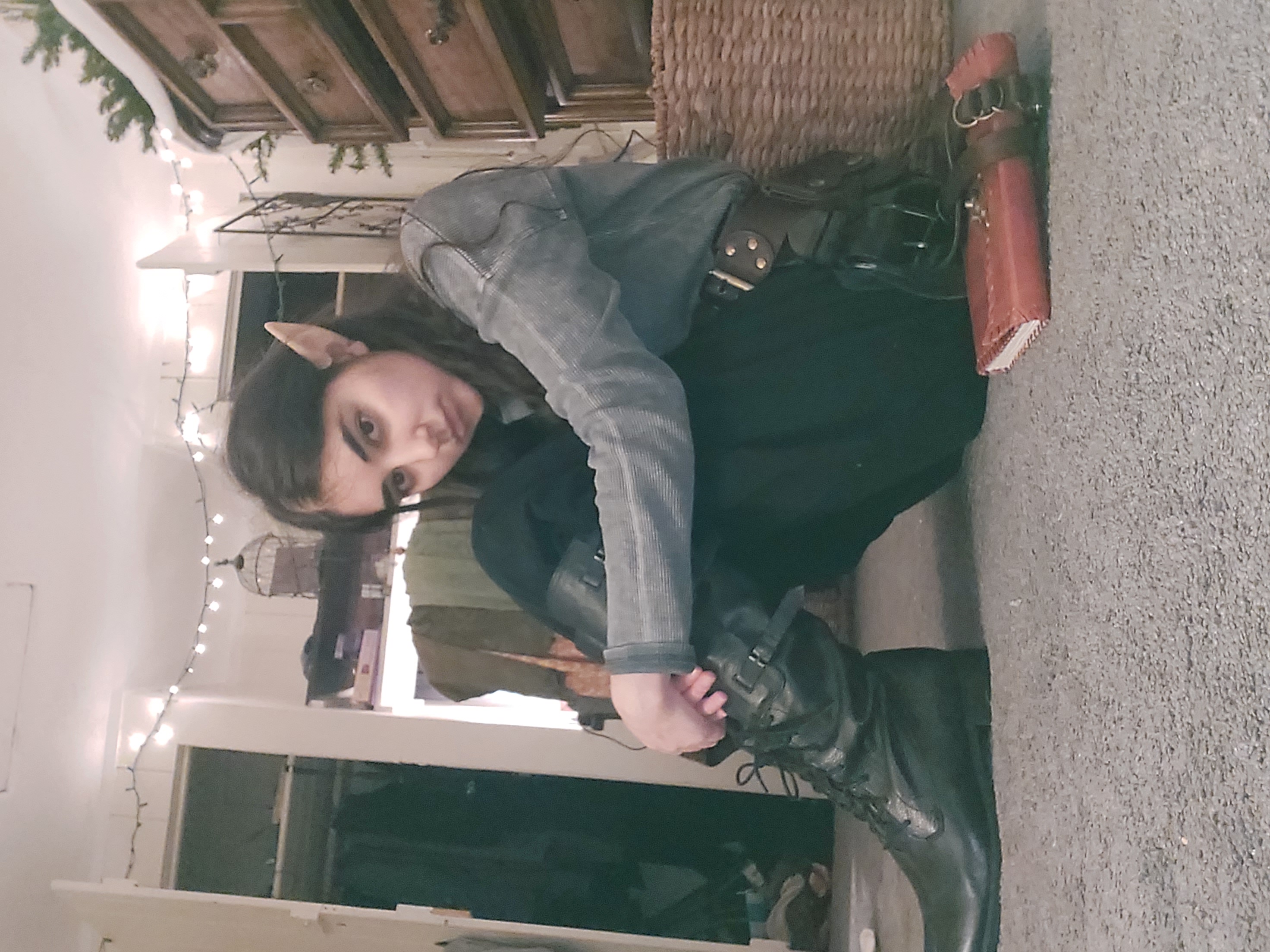 a young woman, possibly a teenager or young adult, is cosplaying as an elf. she is sitting on the floor of a bedroom, wearing elf ears, a grey jacket, dark pants, and tall black leather boots. she has a leather belt with a pouch or holster attached.  a brown leather journal or diary lies on the floor next to her. the room is decorated with string lights.  a wooden dresser and wicker basket are visible in the background.